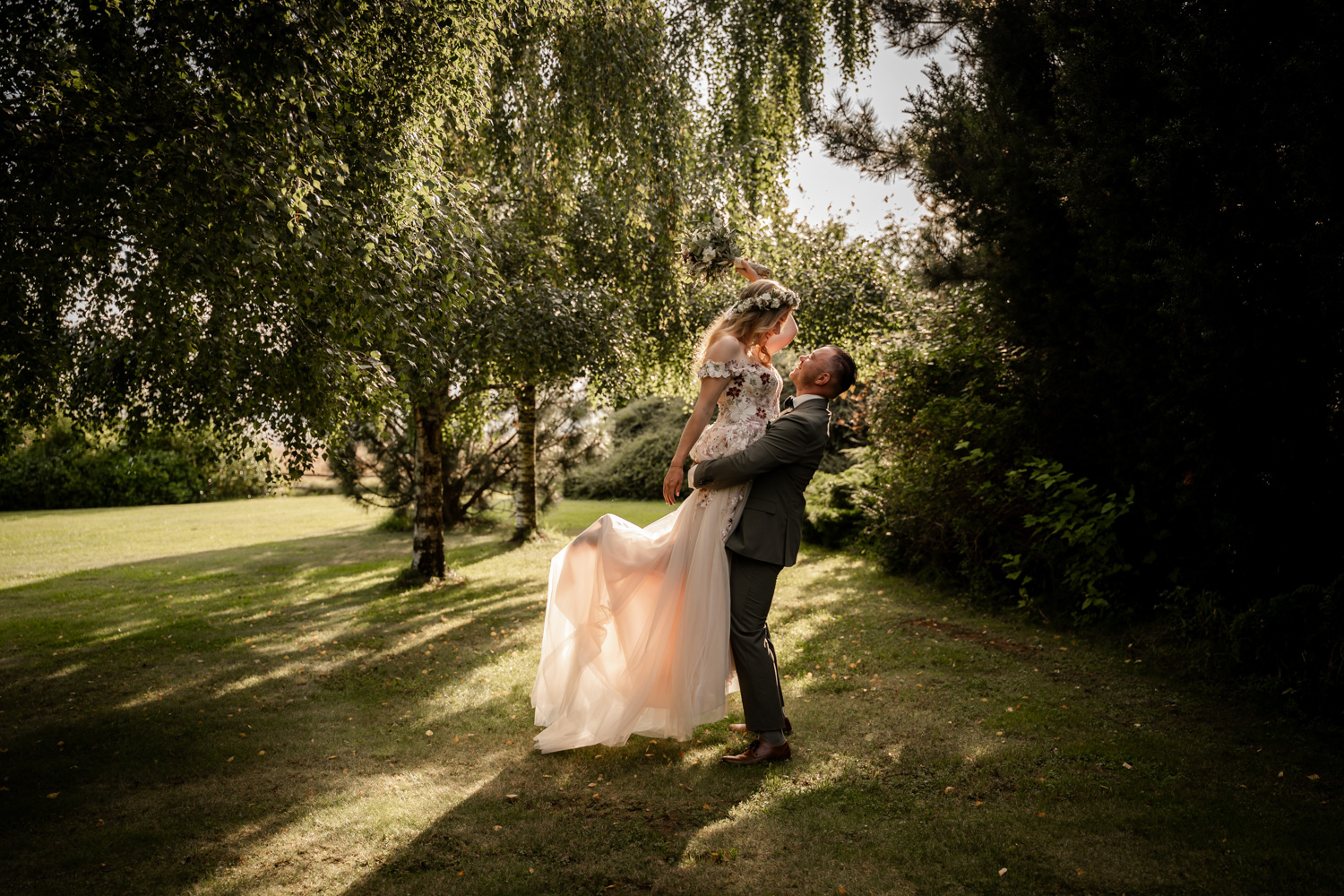 Der Mann trägt seine Braut auf den Armen. Die Sonne leuchtet durchs Kleid. Fotografiert von Anke Hartl von Echte Hochzeitsmomente